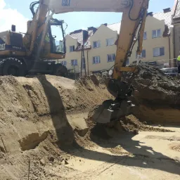 Żółty koparko-ładowarka CAT M315 wykonuje wykop w piaszczystym gruncie, w tle zabudowa miejska i pracownik w odblaskowej kamizelce.