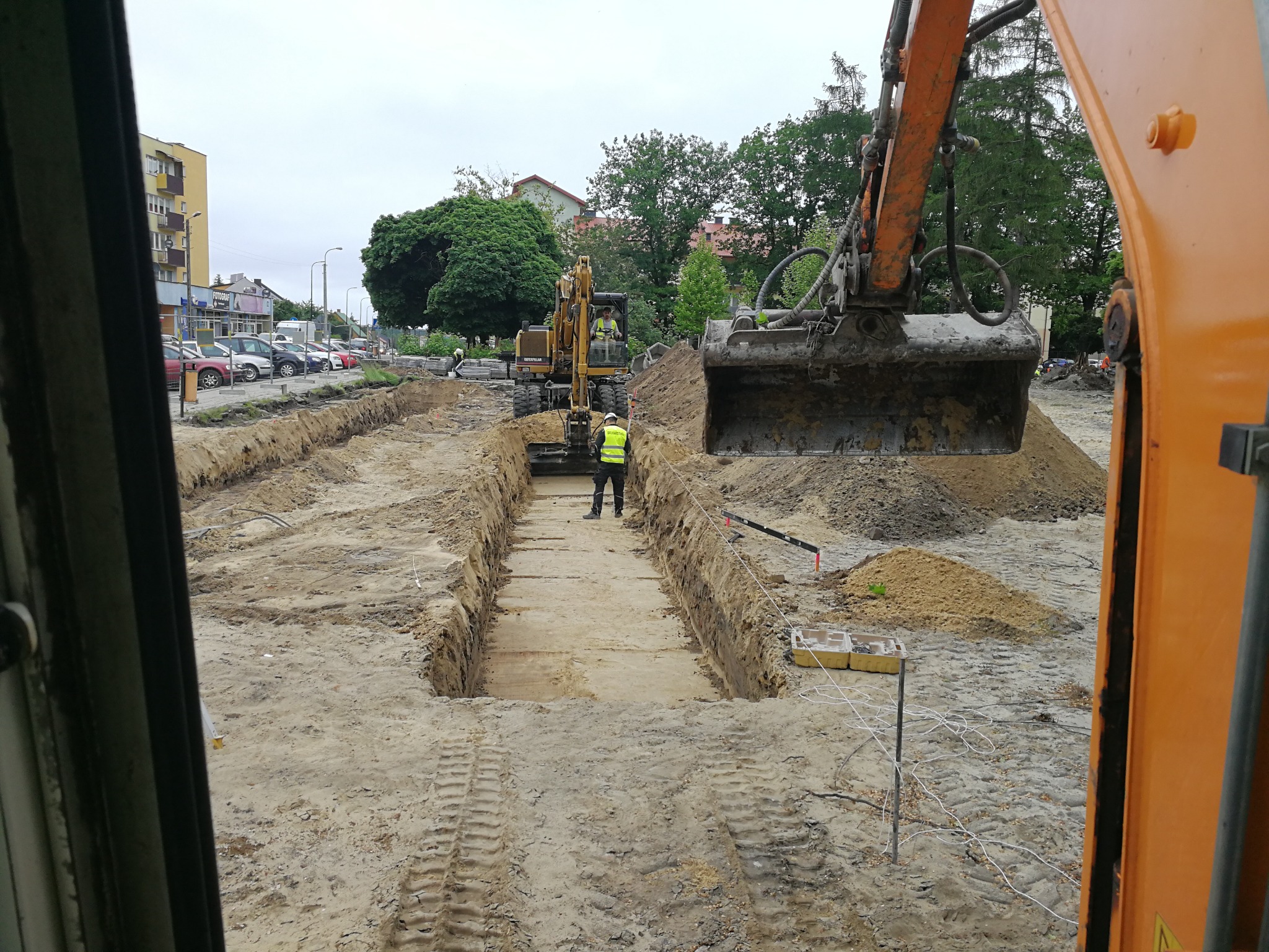 Głęboki wykop liniowy pod instalacje, widok z kabiny koparki, z pracownikiem w kamizelce odblaskowej i drugą koparką w tle.