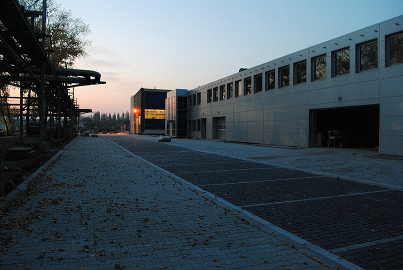 Nowa nawierzchnia z kostki brukowej na parkingu przemysłowym, widoczne jesienne liście i elementy konstrukcyjne w tle, oświetlone wczesnym porankiem.