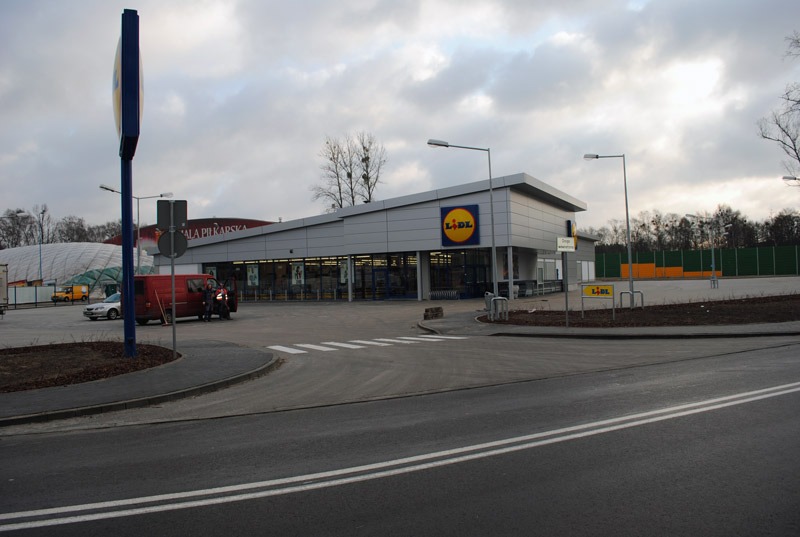 Widok zewnętrzny sklepu Lidl z parkingu i ulicy w pochmurny dzień. Na parkingu zaparkowane samochody i furgonetka.