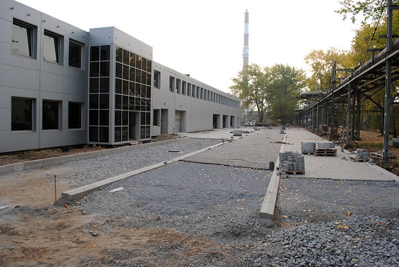 Plac przed nowoczesnym budynkiem w trakcie prac przygotowawczych pod ułożenie nawierzchni z kostki. Widoczne obrzeża, podsypka żwirowa i palety z kostką brukową. W tle komin przemysłowy...