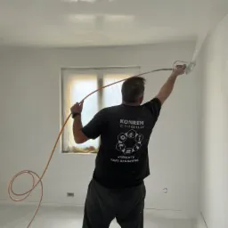 Malowanie ścian natryskowo w pomieszczeniu z oknem zabezpieczonym folią, widoczny pracownik w czarnej koszulce z logo firmy KONREM.