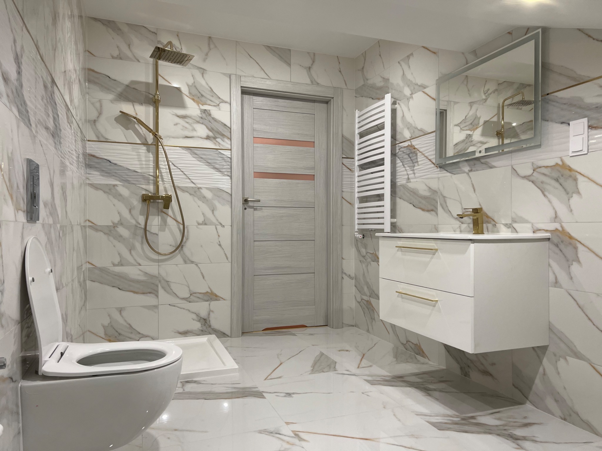 Nowoczesna łazienka z marmurowymi płytkami, złotą armaturą i podwieszaną szafką z umywalką, widok na otwartą toaletę i grzejnik drabinkowy.