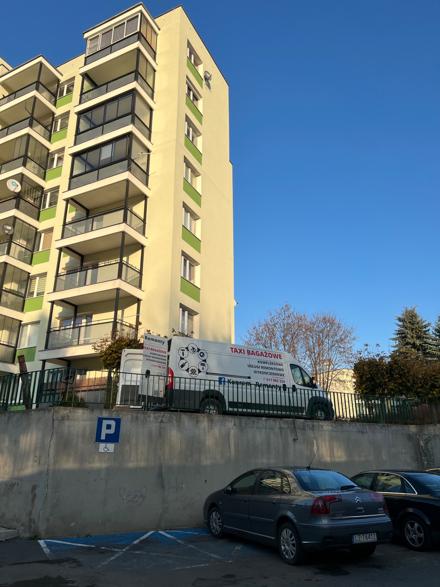 Budynek mieszkalny z balkonami, na parkingu stoi biały bus z napisem 'Taxi Bagażowe - Kompleksowe Usługi Remontowo Wykończeniowe', zaparkowane samochody na pierwszym planie.