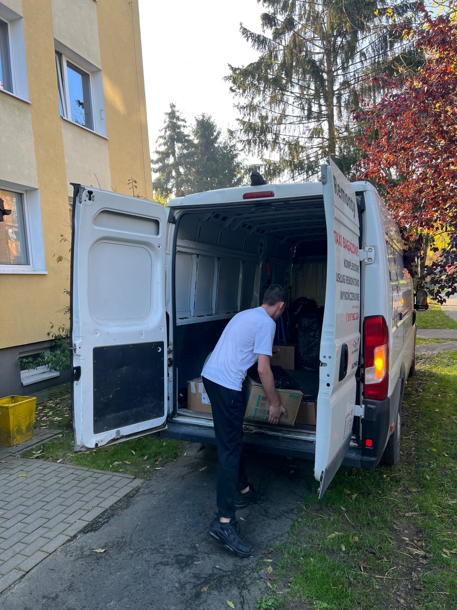 Mężczyzna w białej koszulce wkłada kartonowe pudła do białego busa z otwartymi drzwiami, zaparkowanego na chodniku przed budynkiem, widoczny napis 'Remonty' na karoserii.