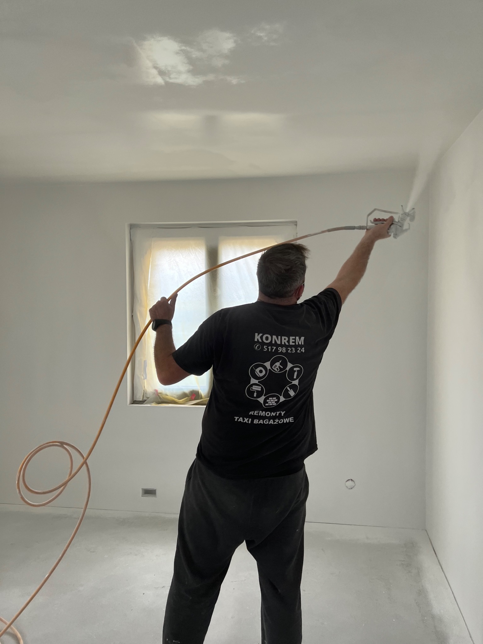 Malowanie ścian natryskowo w pomieszczeniu z oknem zabezpieczonym folią, widoczny pracownik w czarnej koszulce z logo firmy KONREM.