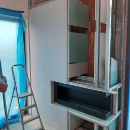 BUDMAR Rosiński Marek - Konstrukcja z płyt karton gips i metalowych profili tworząca obudowę kominka z wnękami, widoczna drabina aluminiowa i zabezpieczona folią okienna wnęka.
