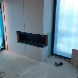 BUDMAR Rosiński Marek - Montaż obudowy kominka z płyt kartonowo-gipsowych, widoczne wkręty, szpachlowanie i narzędzia na podłodze, okno zabezpieczone folią.