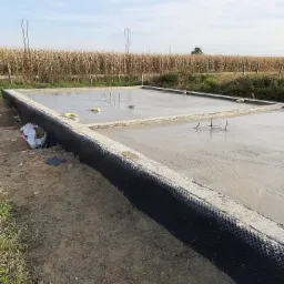 Świeżo wylany fundament z hydroizolacją, wystające pręty zbrojeniowe, w tle pole kukurydzy.