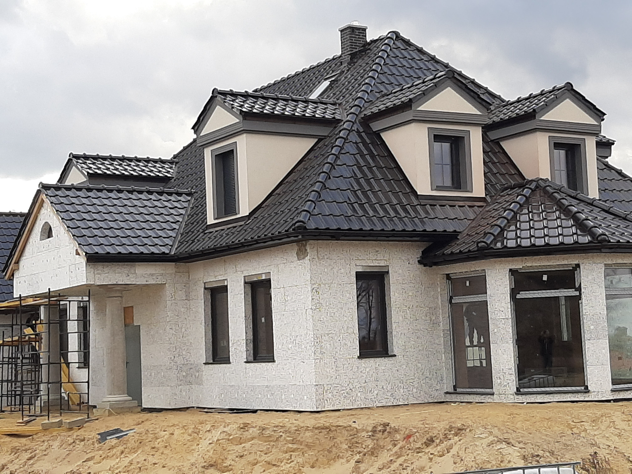 Niedokończony dom jednorodzinny z ciemną dachówką, widoczne rusztowania i piasek wokół budynku.
