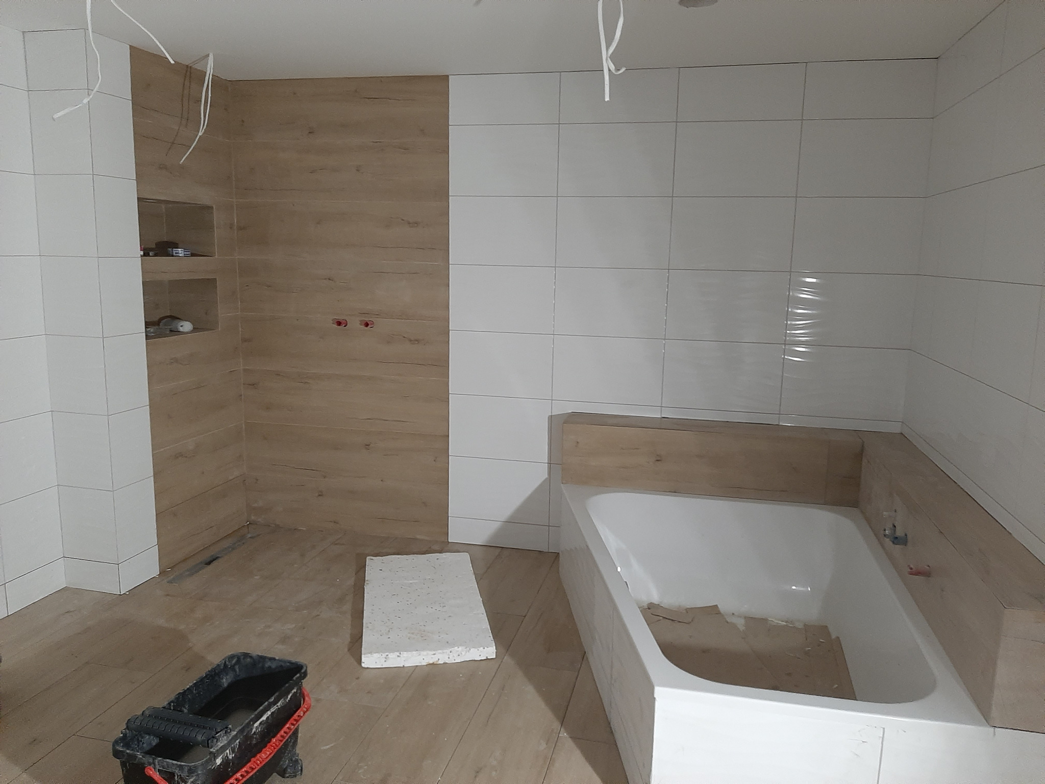Łazienka w trakcie wykańczania: białe i drewnopodobne płytki na ścianach, wanna obudowana płytkami, wiadro z narzędziami na podłodze, widoczne kable zwisające z sufitu.