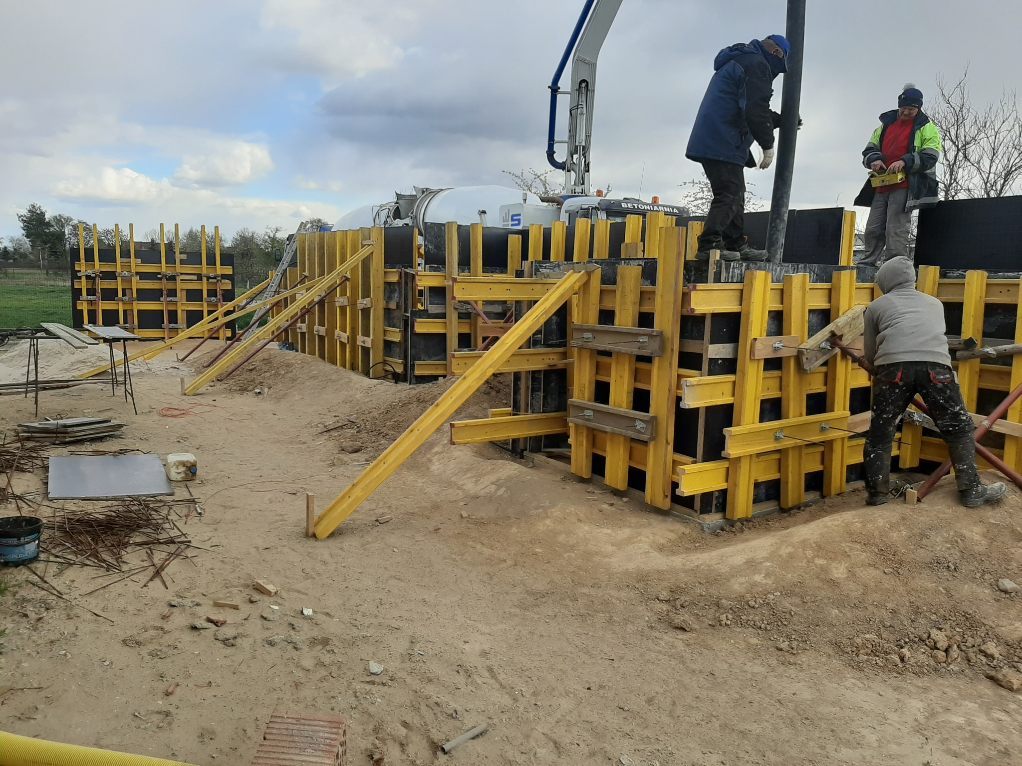 Budowa fundamentów w Pogorzeli: Żółte szalunki zbrojeniowe, robotnicy przy pracy, na drugim planie betonowóz z pompą do betonu.