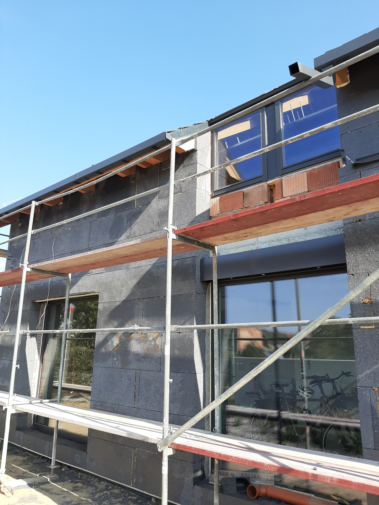 Ściana zewnętrzna domu w trakcie ocieplania styropianem grafitowym, widoczne rusztowanie, okna z roletami zewnętrznymi i częściowo wymurowana nadproża z czerwonej cegły.