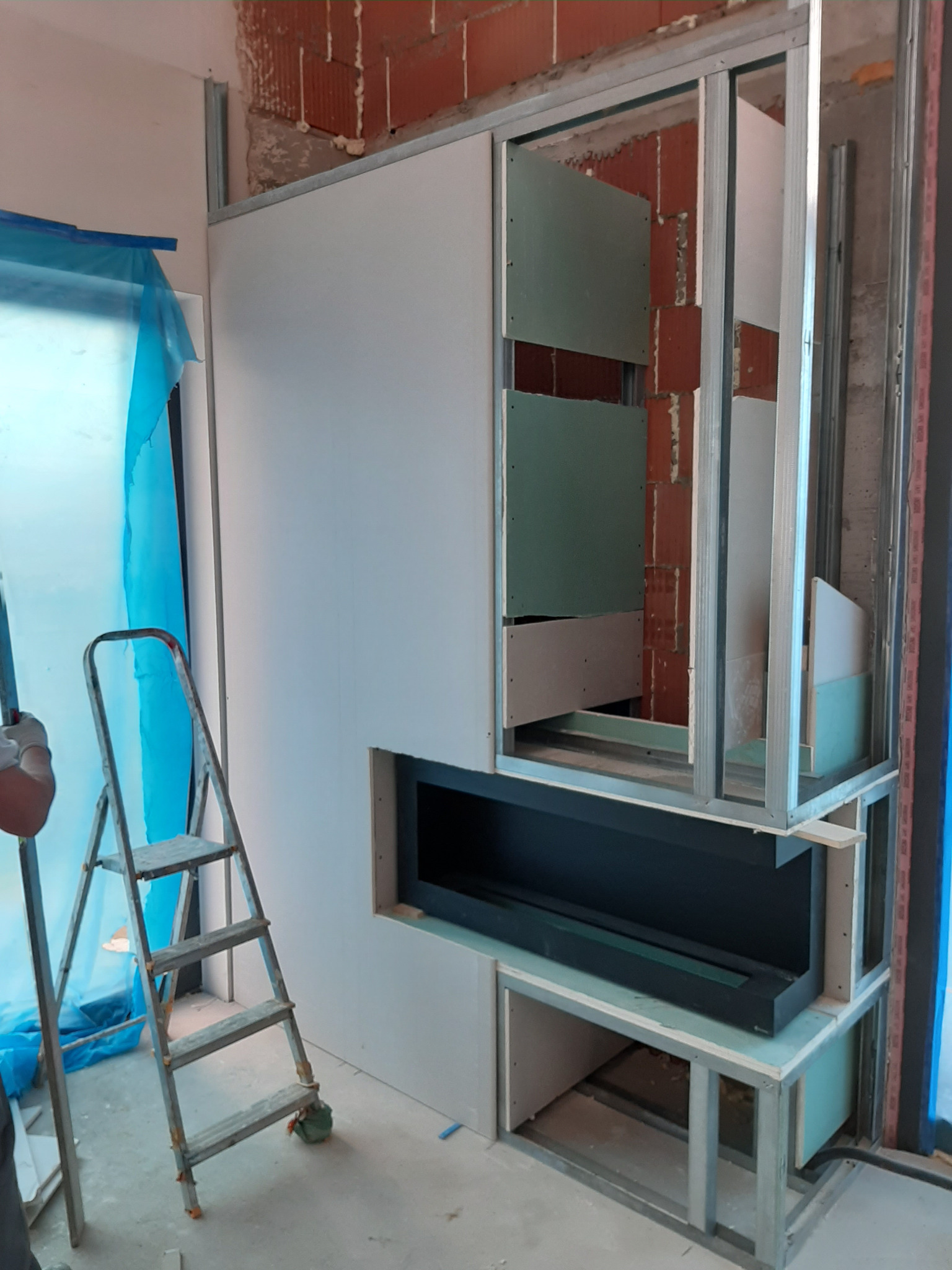 Konstrukcja z płyt karton gips i metalowych profili tworząca obudowę kominka z wnękami, widoczna drabina aluminiowa i zabezpieczona folią okienna wnęka.