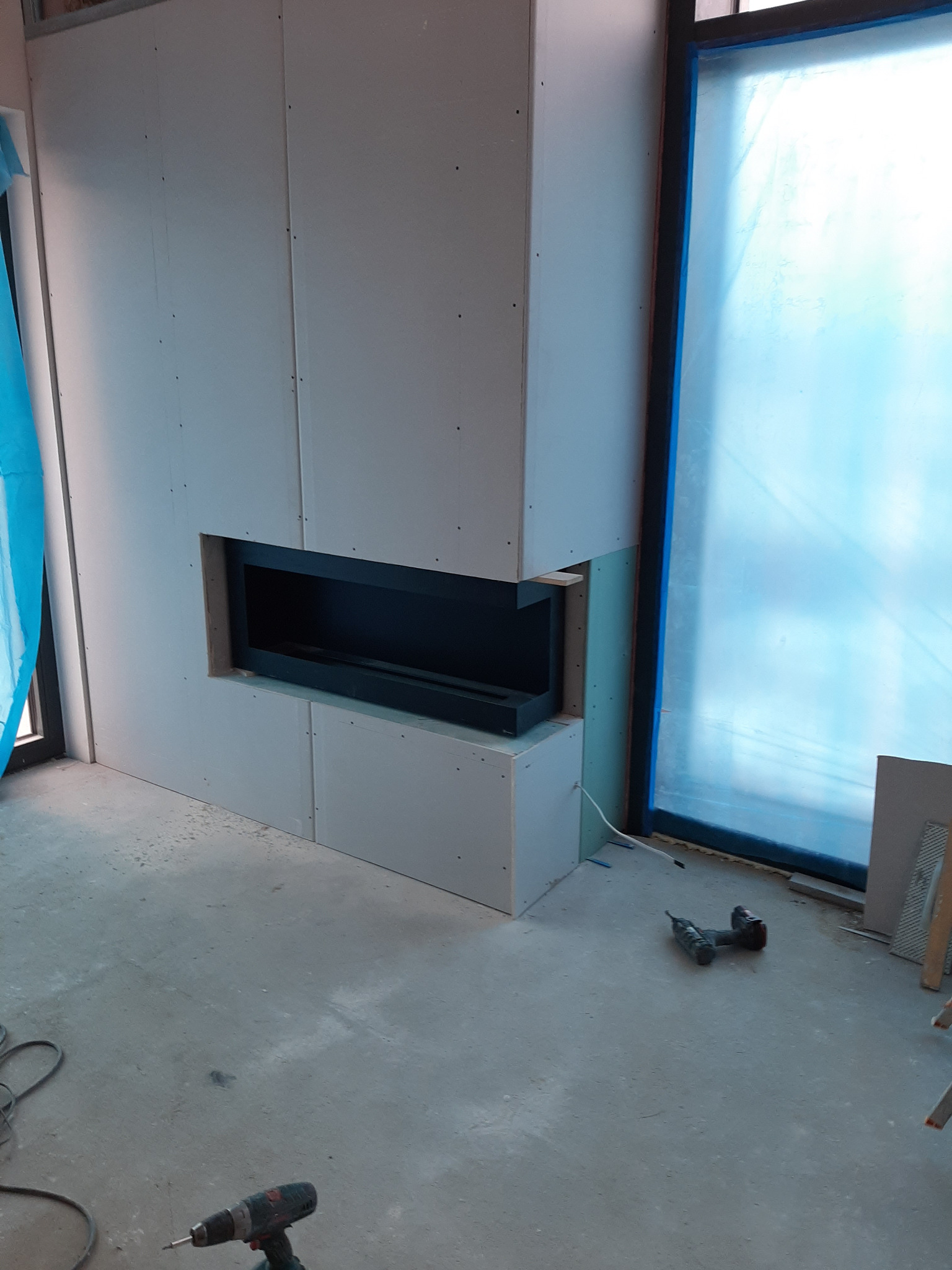 Montaż obudowy kominka z płyt kartonowo-gipsowych, widoczne wkręty, szpachlowanie i narzędzia na podłodze, okno zabezpieczone folią.