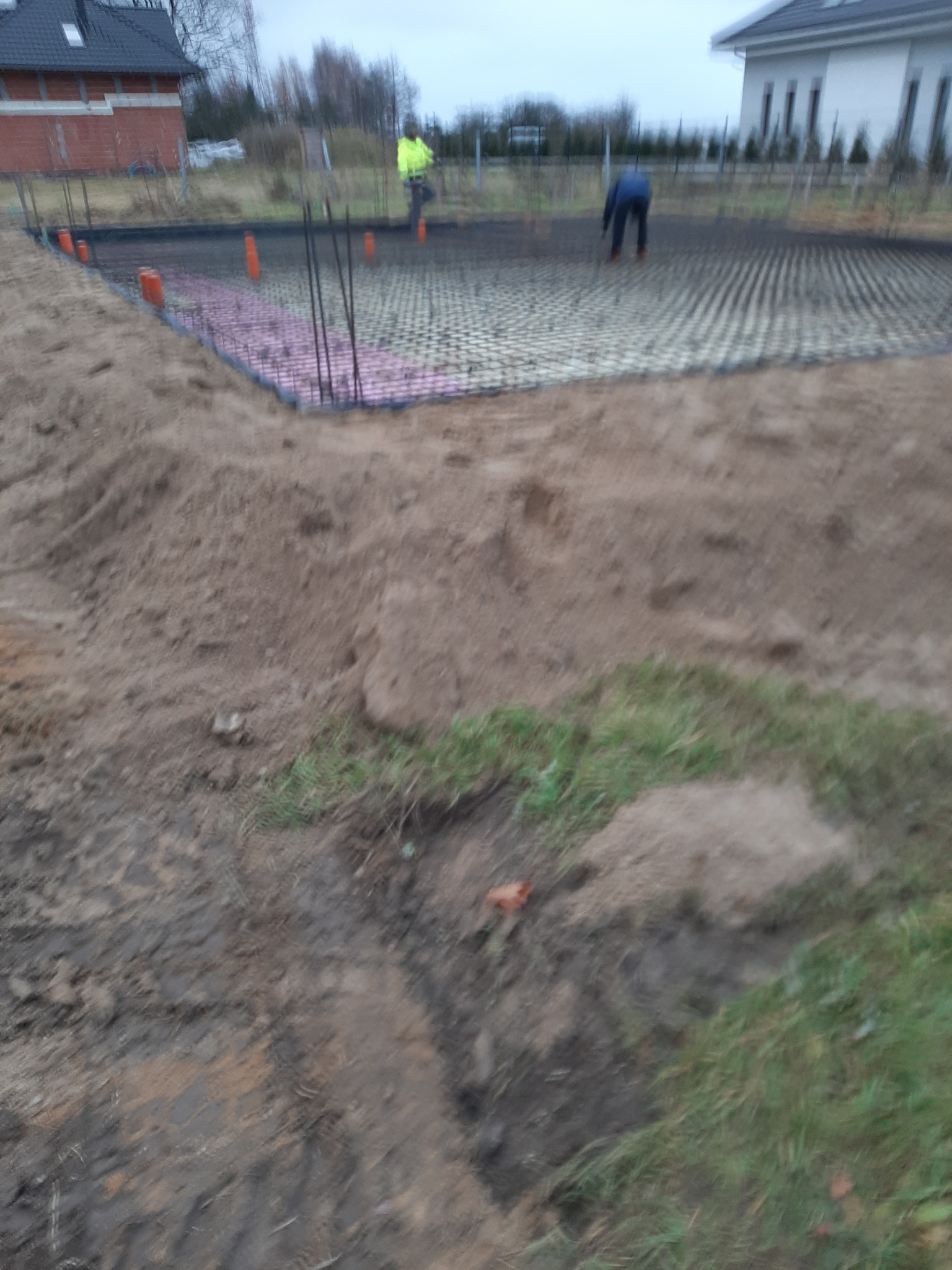 Przygotowanie terenu pod budowę domu: widoczne fundamenty z szalunkiem, zbrojenie pionowe, warstwa izolacyjna z różowego styropianu, pracownicy w tle.