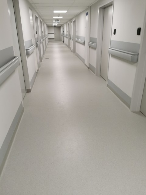 Długi korytarz w placówce medycznej z jasną, jednolitą podłogą winylową, zabezpieczony odbojnicami ściennymi i listwami przypodłogowymi w kolorze szarym.