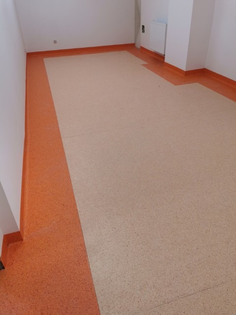 Przygotowanie do montażu wykładziny: podkład korkowy rozłożony na pomarańczowej wykładzinie podłogowej w pustym pomieszczeniu z białymi ścianami i grzejnikiem.