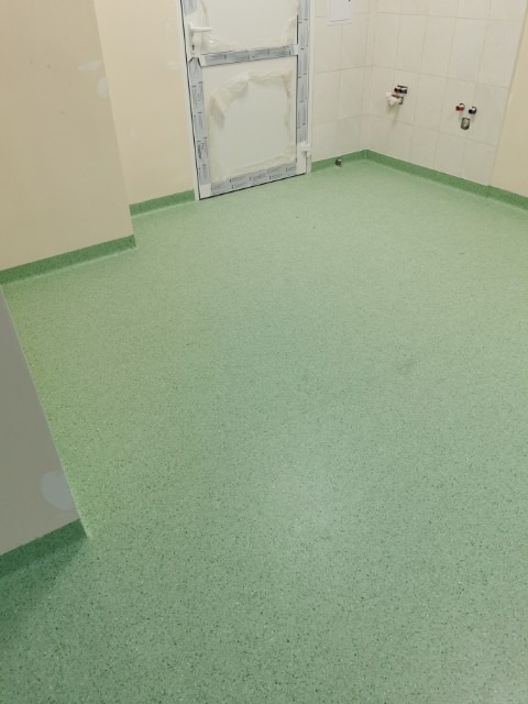 Jasnozielona podłoga z wykładziny PCV w pomieszczeniu z białymi ścianami i drzwiami, widoczne przyłącza wodne na ścianie.