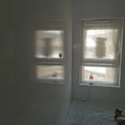 Świeżo pomalowane na biało ściany w pokoju, okno zabezpieczone folią, na parapecie pusta butelka po napoju, podłoga zakryta folią malarską, widoczne narzędzia malarskie.