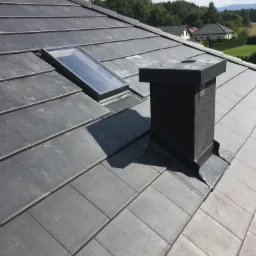 Szary dach z dachówki ceramicznej z zamontowanym oknem dachowym i kominem wentylacyjnym, w tle widok na domy i zieleń.