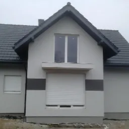 Gotowy dom z szarą dachówką i elewacją w odcieniach bieli i szarości. Widoczne rolety zewnętrzne i fragment terenu przed budynkiem.