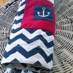 Rolowana, pikowana mata do zabawy w marynarskim stylu: biało-granatowe zygzaki, czerwona kieszeń z kotwicą, tasiemki z motywem kotwicy. Leży na wiklinowym koszu.