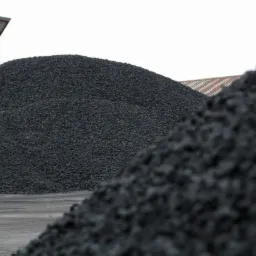 Góra czarnego węgla pod zadaszeniem. Widoczna faktura węgla i fragment budynku w tle. Ujęcie z dołu podkreśla wielkość hałdy.