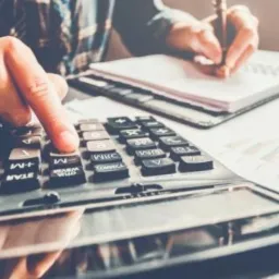 Osoba w koszuli w kratę oblicza coś na kalkulatorze, notując w notatniku, obok leżą dokumenty z wykresami.