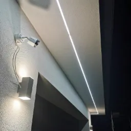 Zewnętrzna kamera monitoringu Hikvision zamontowana na elewacji budynku, obok lampy ściennej, z widocznymi przewodami i oświetleniem LED w suficie podwieszanym.