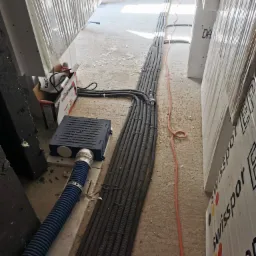 Wiązka kabli elektrycznych ułożona w korycie podłogowym w surowym stanie, widoczna niebieska rura karbowana, puszka instalacyjna i kartonowe pudełka z narzędziami Yato.