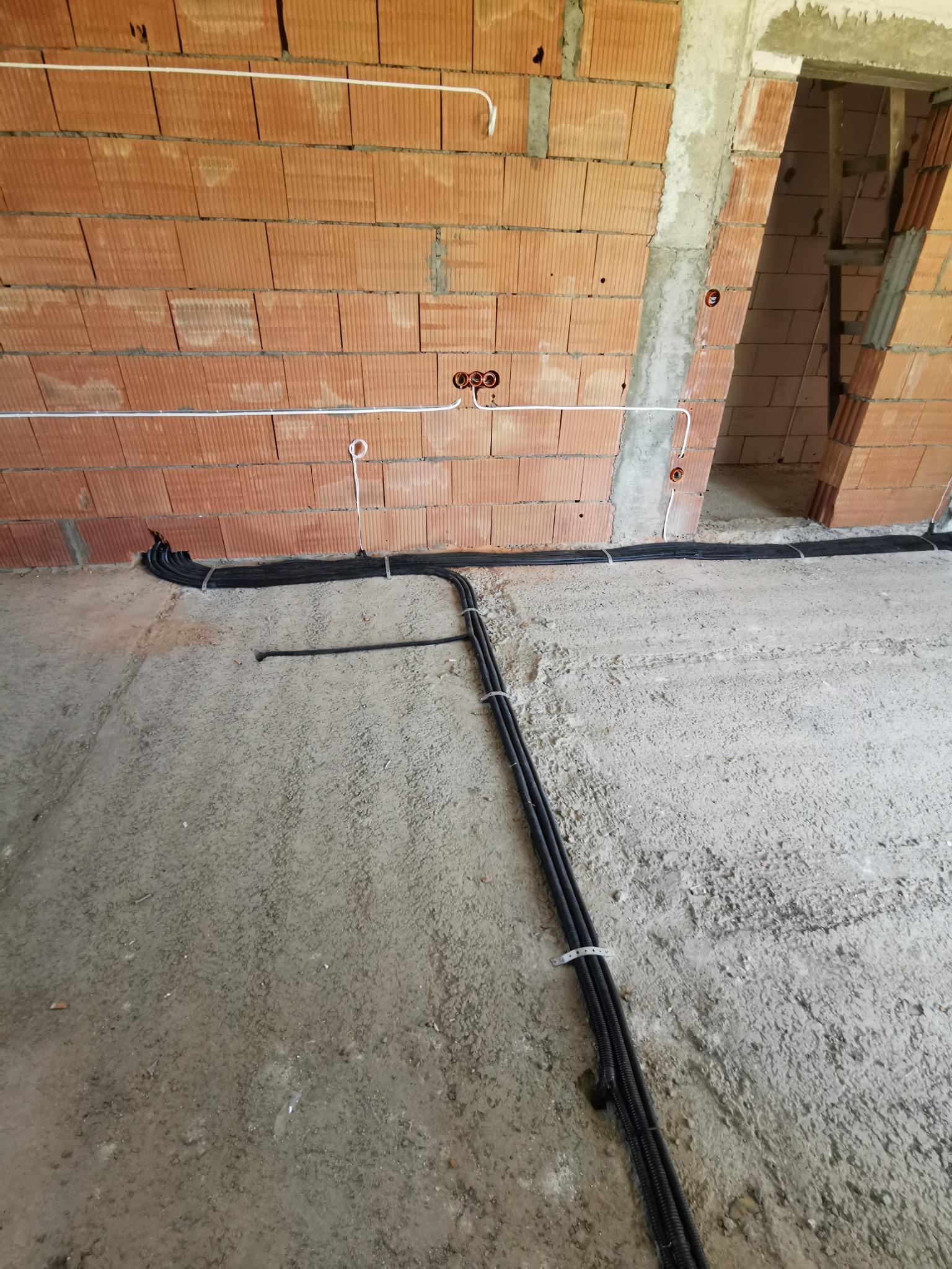 Surowy stan wnętrza budynku w trakcie prac elektrycznych: widoczne kable w peszlach ułożone na szarej podłodze, białe przewody pod sufitem i otwory na gniazdka w ścianie z czerwonej cegły.