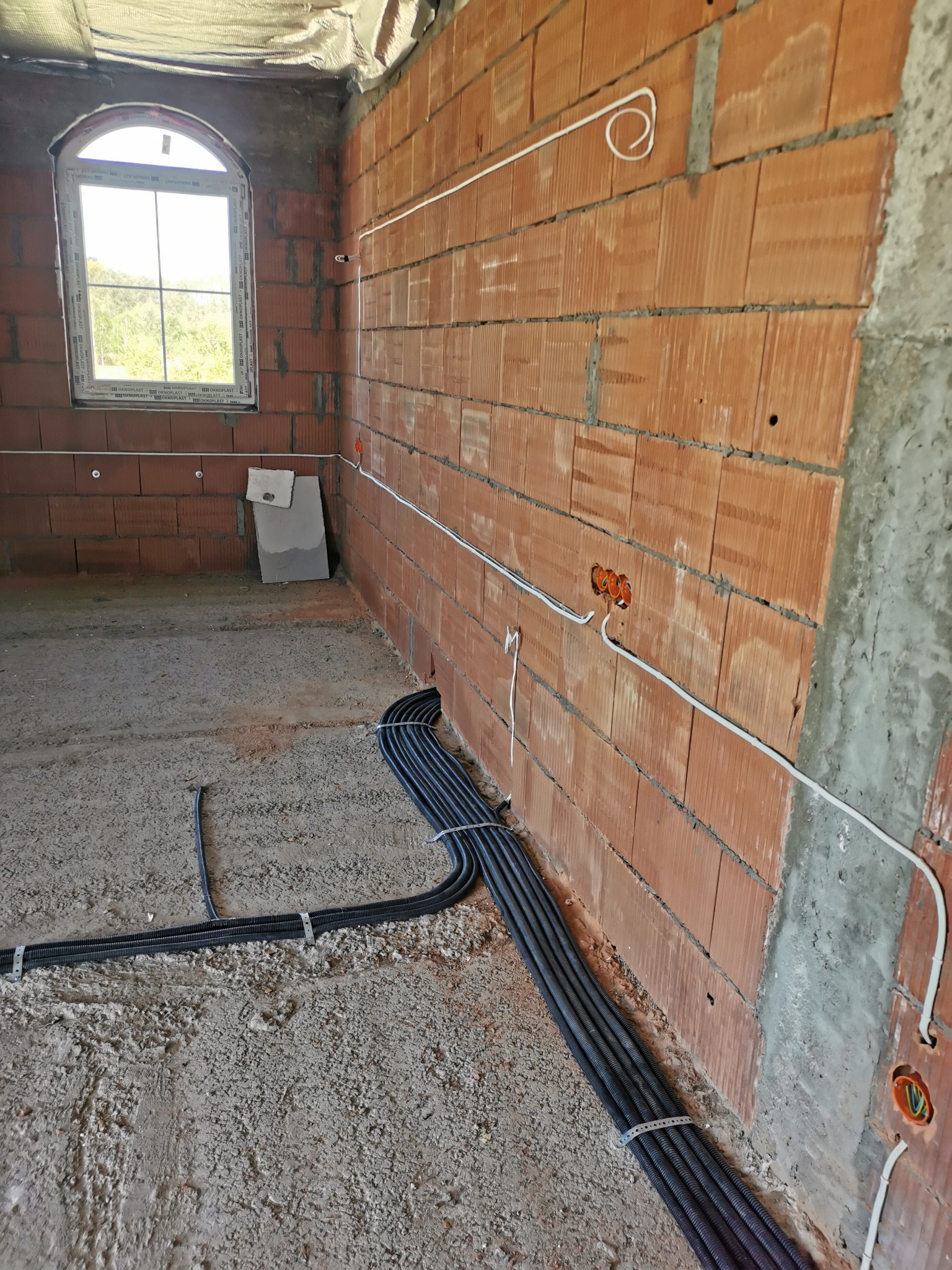 Wnętrze budynku w trakcie instalacji elektrycznej: widoczne kable prowadzone po ścianie z cegły, puszki elektryczne i okno łukowe w stanie surowym.