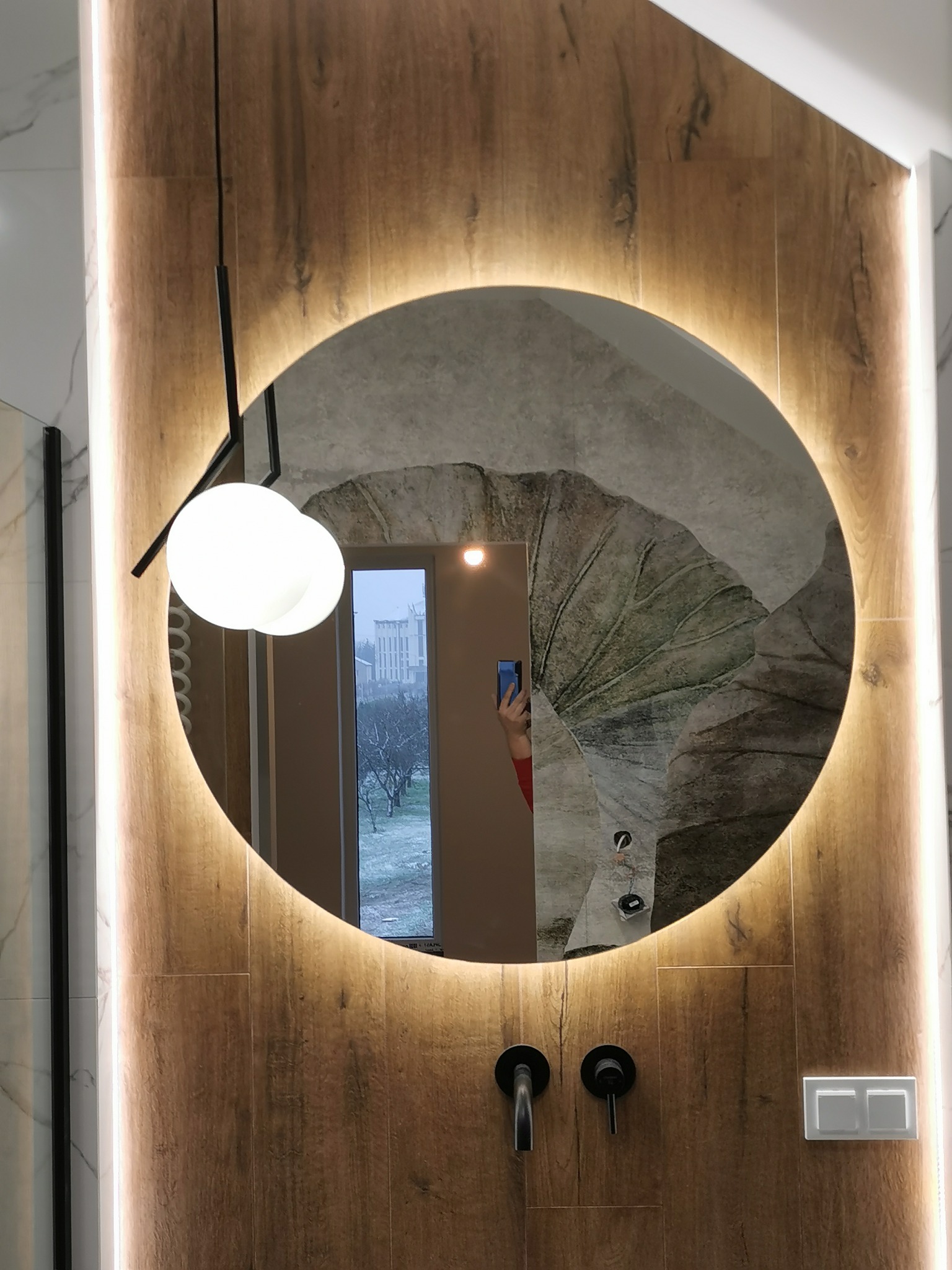 Okrągłe lustro z podświetleniem LED w łazience z drewnopodobnymi panelami, widoczna minimalistyczna lampa wisząca i czarna armatura, odbicie okna z widokiem na zaśnieżony krajobraz.