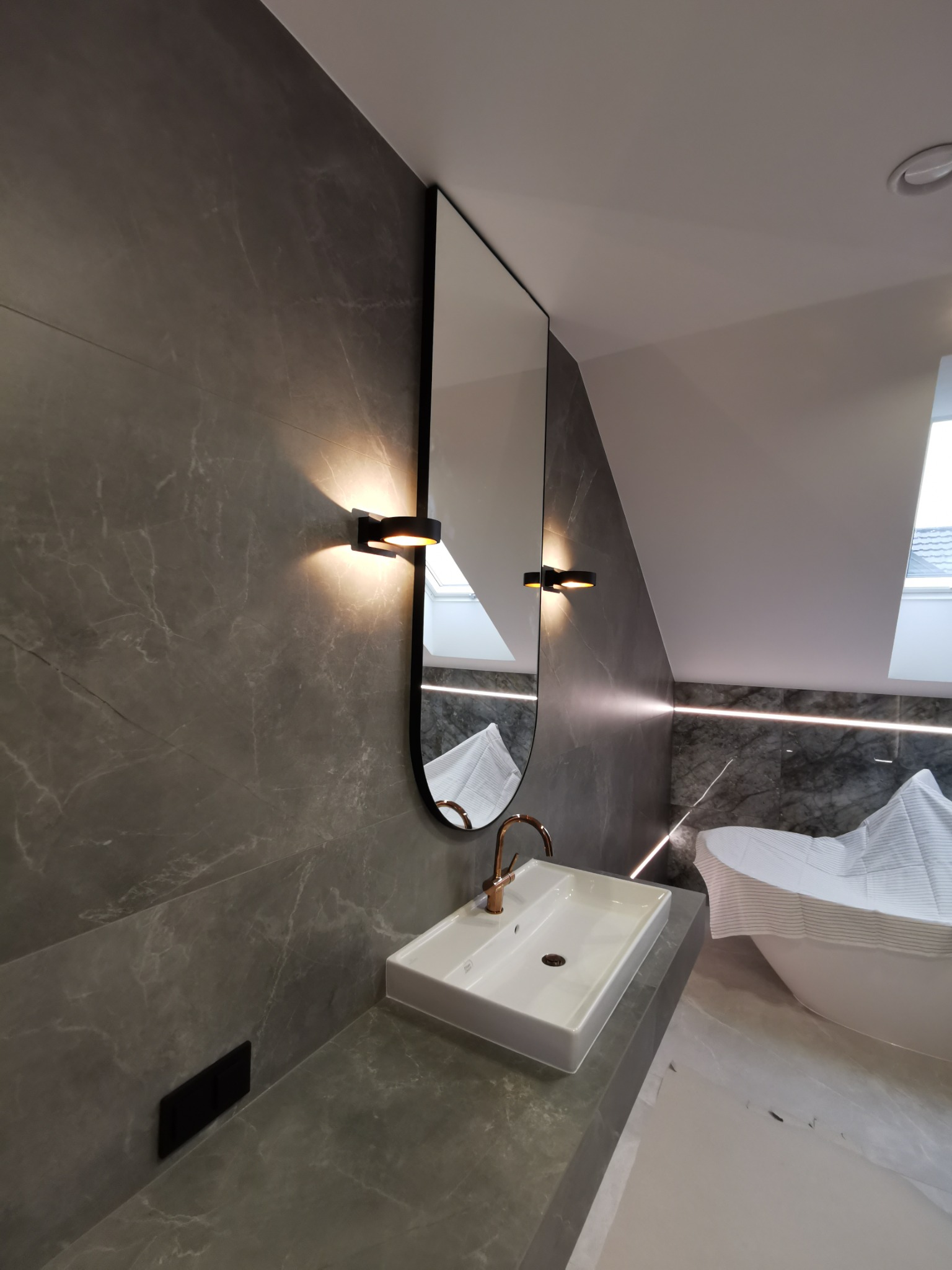 Nowoczesna łazienka na poddaszu z umywalką nablatową, złotą baterią i podświetlanym lustrem owalnym oraz szarą marmurową ścianą i ukrytym włącznikiem światła.