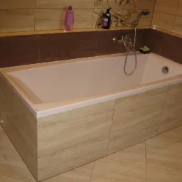 Wykończona wanna obudowana płytkami imitującymi drewno, z baterią i słuchawką prysznicową, na tle płytek w odcieniach beżu i brązu, z kosmetykami na półce.