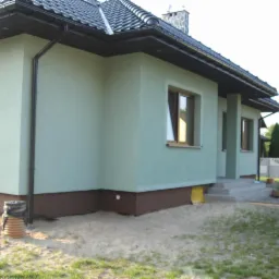 Świeżo otynkowany dom jednorodzinny z brązowym cokołem i grafitową dach&oacute;wką, widoczny z boku, z fragmentem trawnika i piasku przed budynkiem.