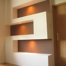 Narożna zabudowa z płyt gipsowo-kartonowych tworząca dekoracyjne wnęki z oświetleniem punktowym, pomalowane na biało i brązowo, sąsiadująca z drewnianymi drzwiami.