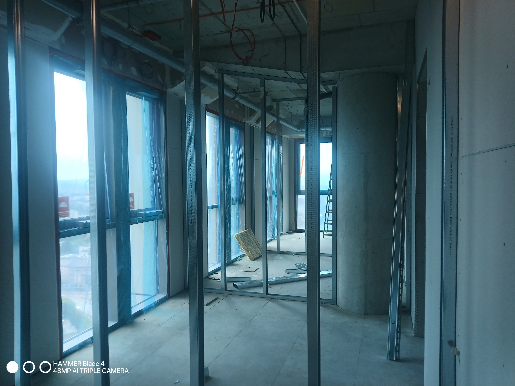 Wnętrze w trakcie prac: metalowa konstrukcja szkieletowa pod zabudowę ścian działowych z płyty gipsowo-kartonowej, okna zabezpieczone folią, widoczne materiały izolacyjne.