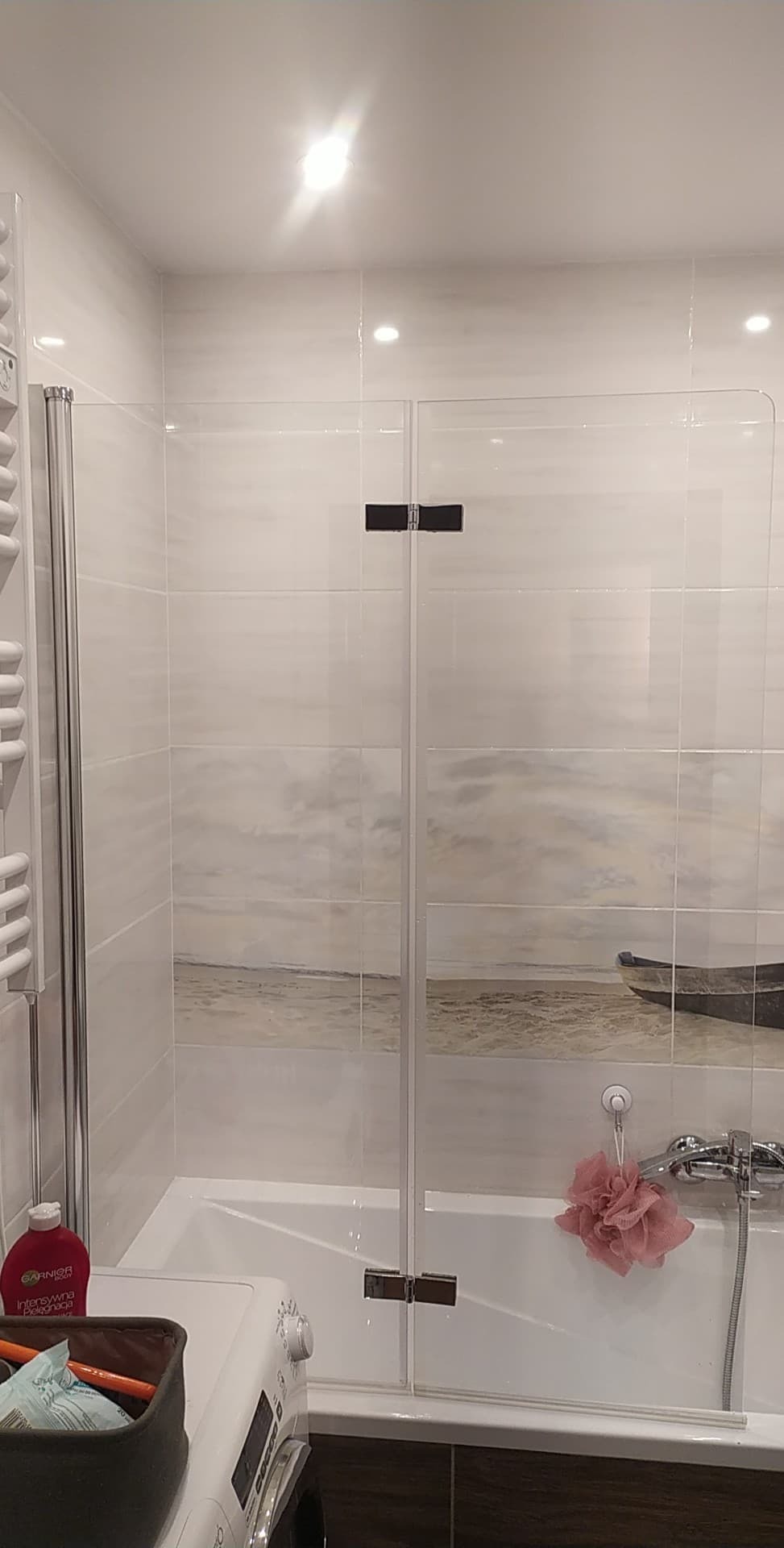 Szklana kabina nawannowa z czarnymi zawiasami, zamontowana na wannie z panelem dekoracyjnym imitującym plażę i łódź, widoczna pralka i akcesoria łazienkowe.