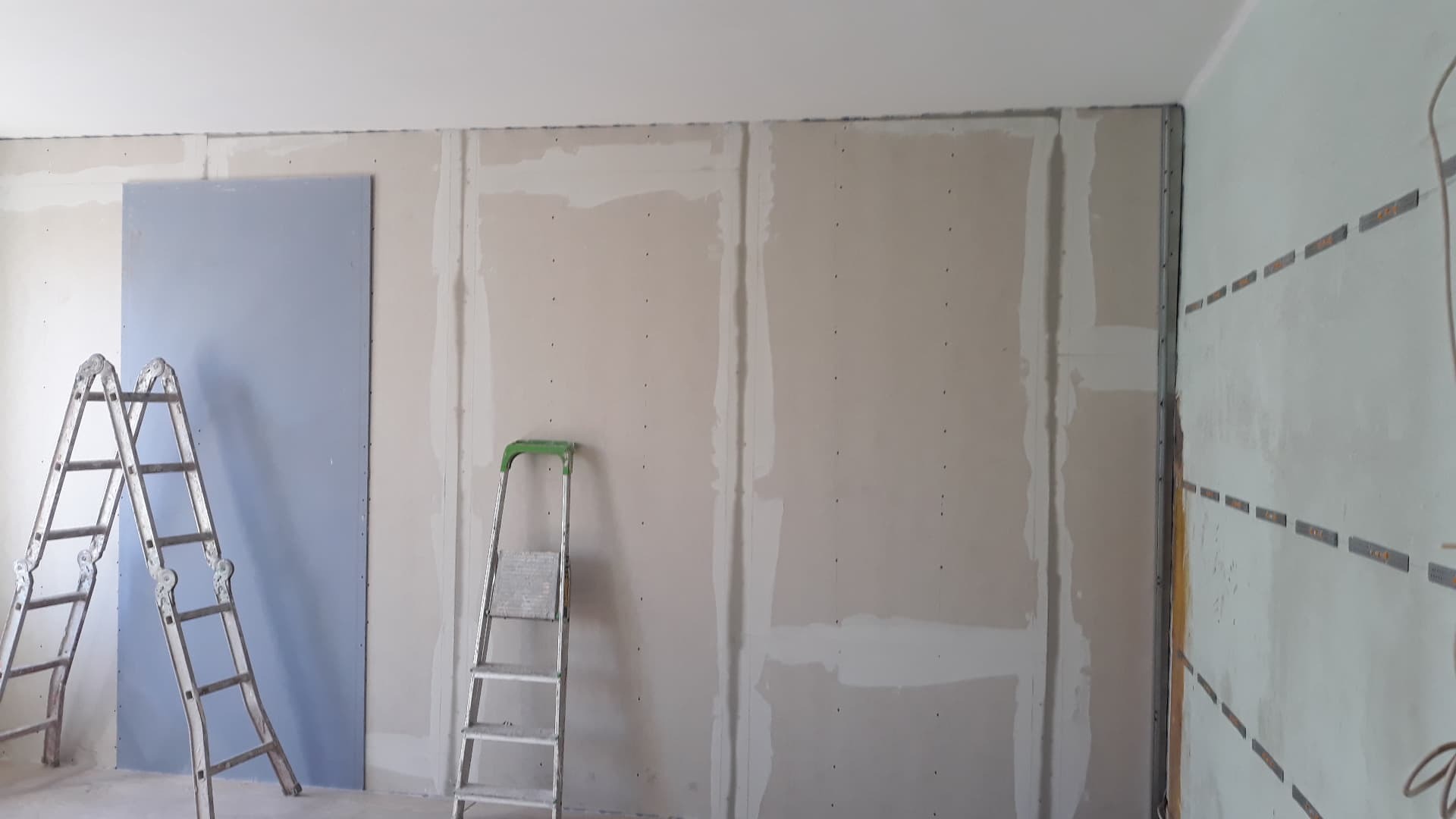 Montaż płyt gipsowo-kartonowych na ścianie z widocznymi profilami, dwie drabiny aluminiowe w pomieszczeniu w trakcie prac.