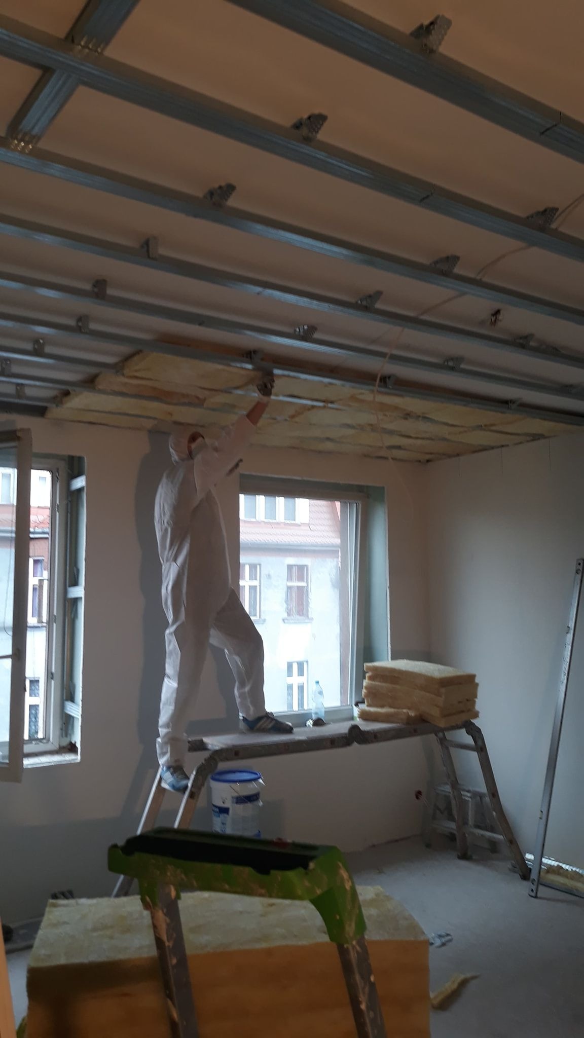 Pracownik w kombinezonie montuje izolację z wełny mineralnej w suficie podwieszanym na metalowej konstrukcji w mieszkaniu w Tychach, widok z dołu na drabinę i okno.
