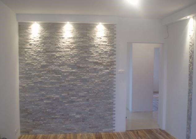 Ściana wykończona szarą okładziną kamienną, oświetlona trzema punktowymi lampami sufitowymi, widok na wnętrze przez otwarte drzwi.