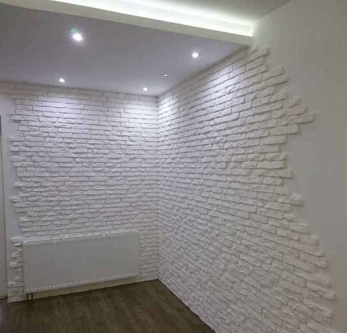Fragment ściany wykończonej białym kamieniem dekoracyjnym, ułożonym w nieregularny wzór cegiełek, z widocznym grzejnikiem panelowym i oświetleniem sufitowym LED.
