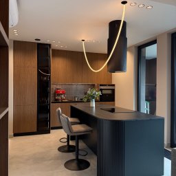 Damko Damian Kozłowski - Nowoczesna kuchnia z wyspą w kolorze grafitu, drewnianymi szafkami i oryginalnym oświetleniem. Widoczne krzesła barowe i ekspres do kawy.