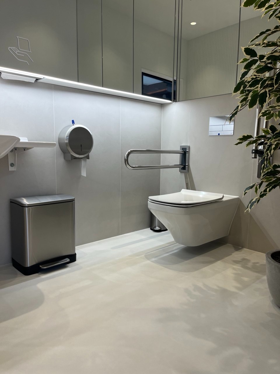 Nowoczesna toaleta dla niepełnosprawnych z wiszącą muszlą, uchwytem, podajnikiem papieru i koszem. Minimalistyczny design w jasnych szarościach, z rośliną doniczkową.