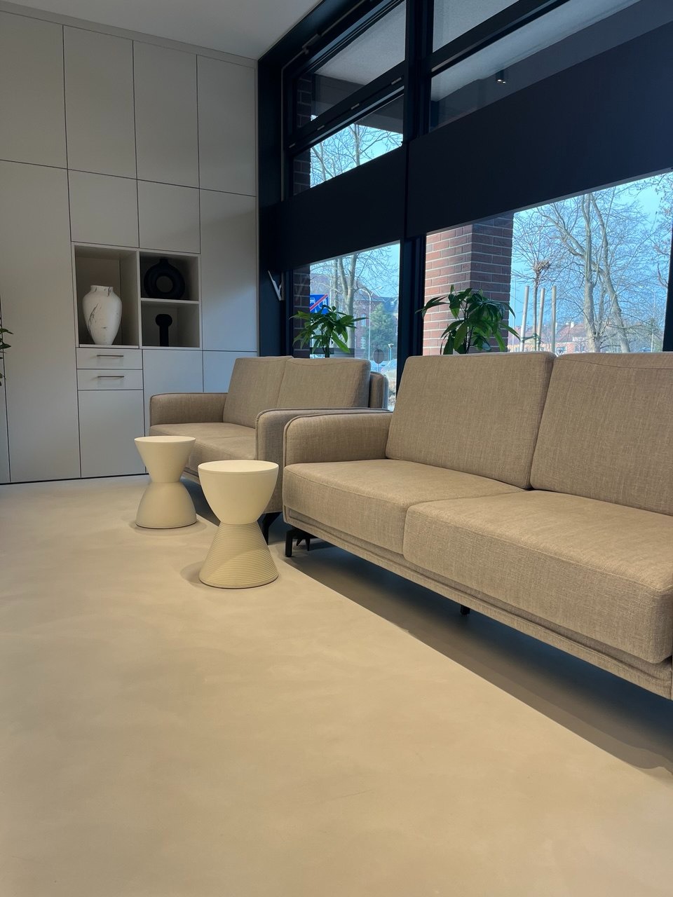 Jasne wnętrze z dwoma sofami i stolikami w kształcie klepsydry, duże okno z widokiem na zewnątrz, minimalistyczna zabudowa ścienna z półkami i dekoracjami.