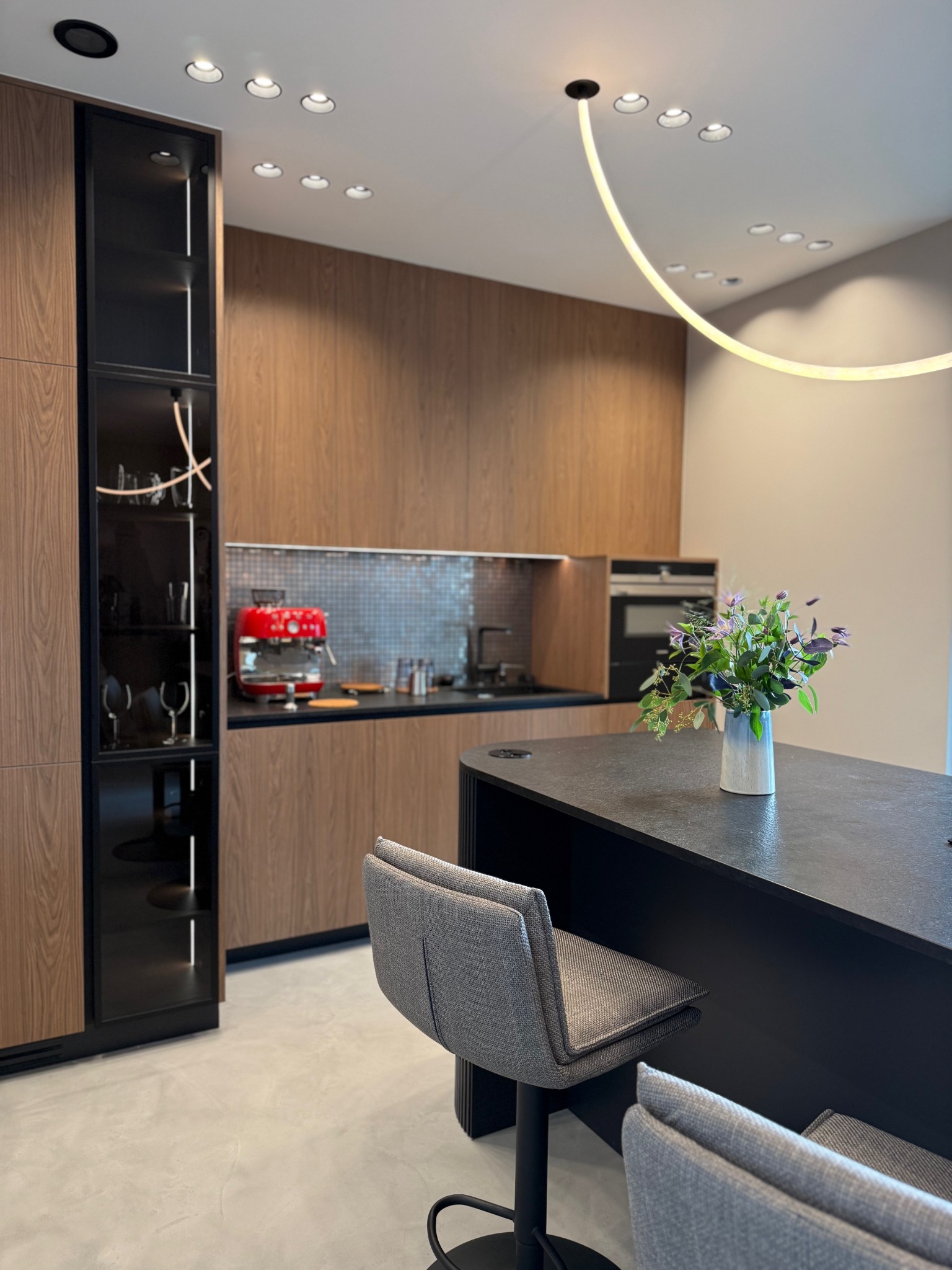 Nowoczesna kuchnia z drewnianymi szafkami, czarnym blatem i wyspą. Na blacie wazon z kwiatami. Oświetlenie punktowe i dekoracyjna lampa łukowa.