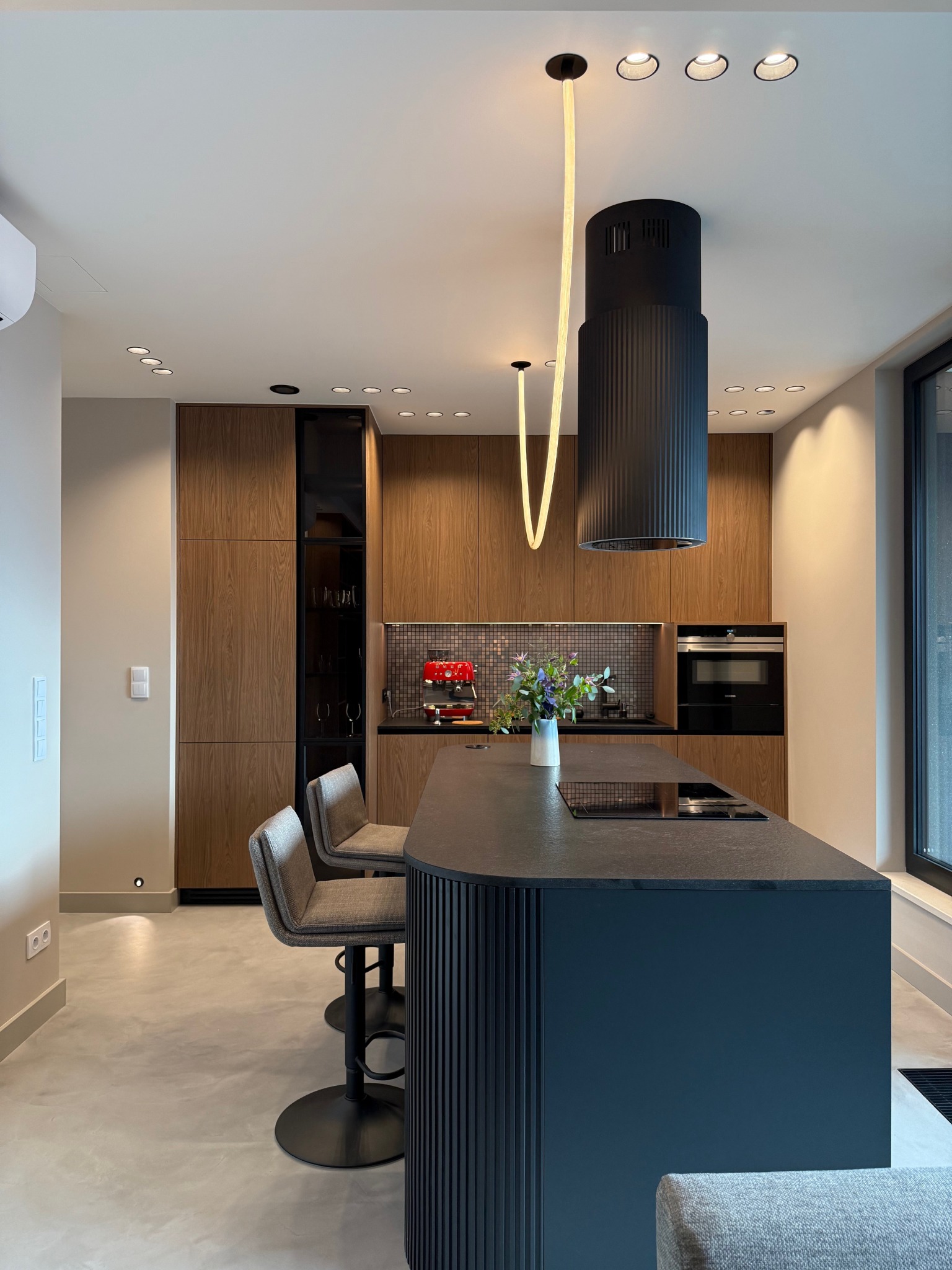 Nowoczesna kuchnia z wyspą, drewnianymi szafkami i designerskim oświetleniem. Minimalistyczny wystrój, czarne akcenty i kwiaty w wazonie na blacie.