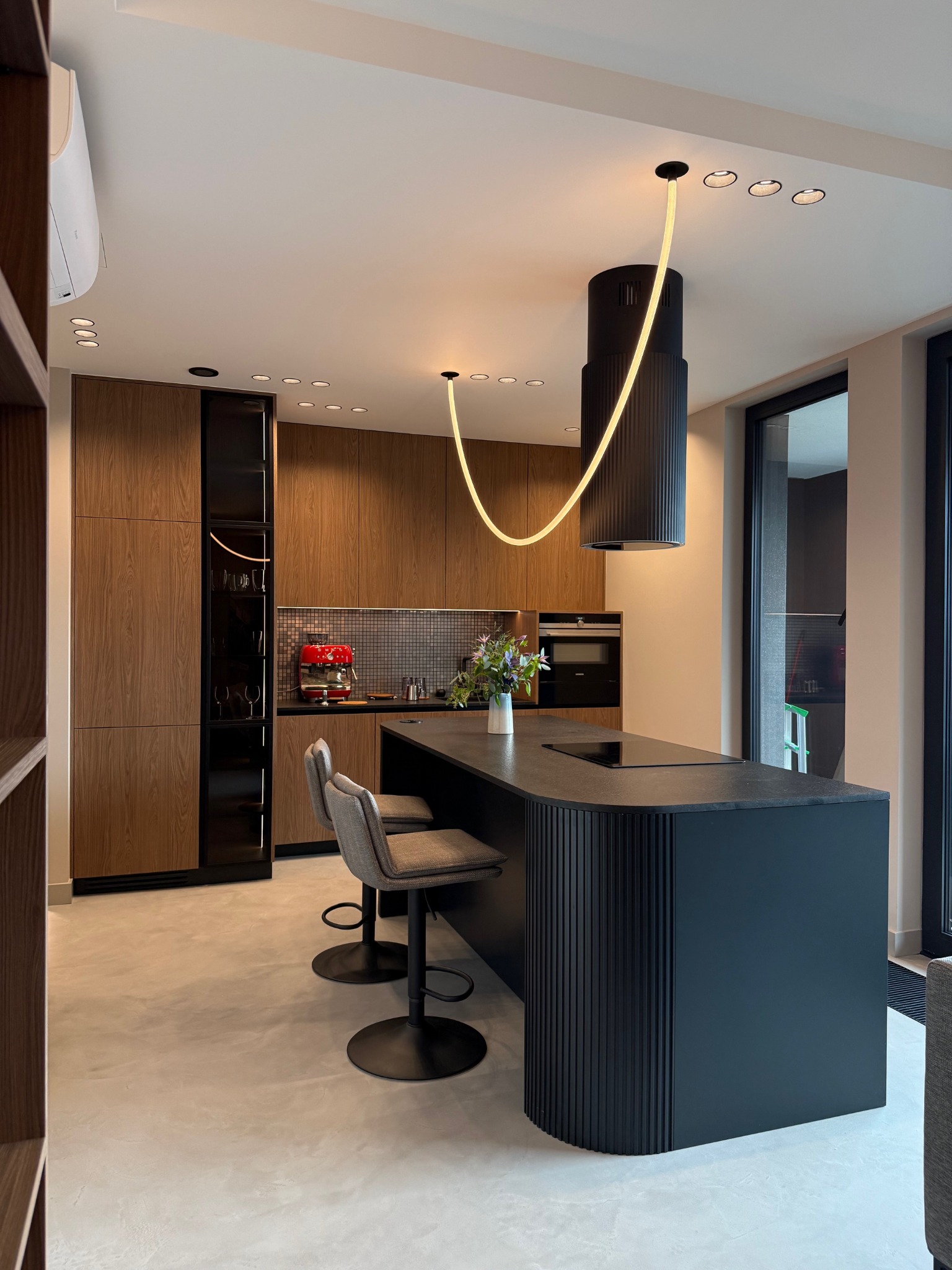 Nowoczesna kuchnia z wyspą w kolorze grafitu, drewnianymi szafkami i oryginalnym oświetleniem. Widoczne krzesła barowe i ekspres do kawy.
